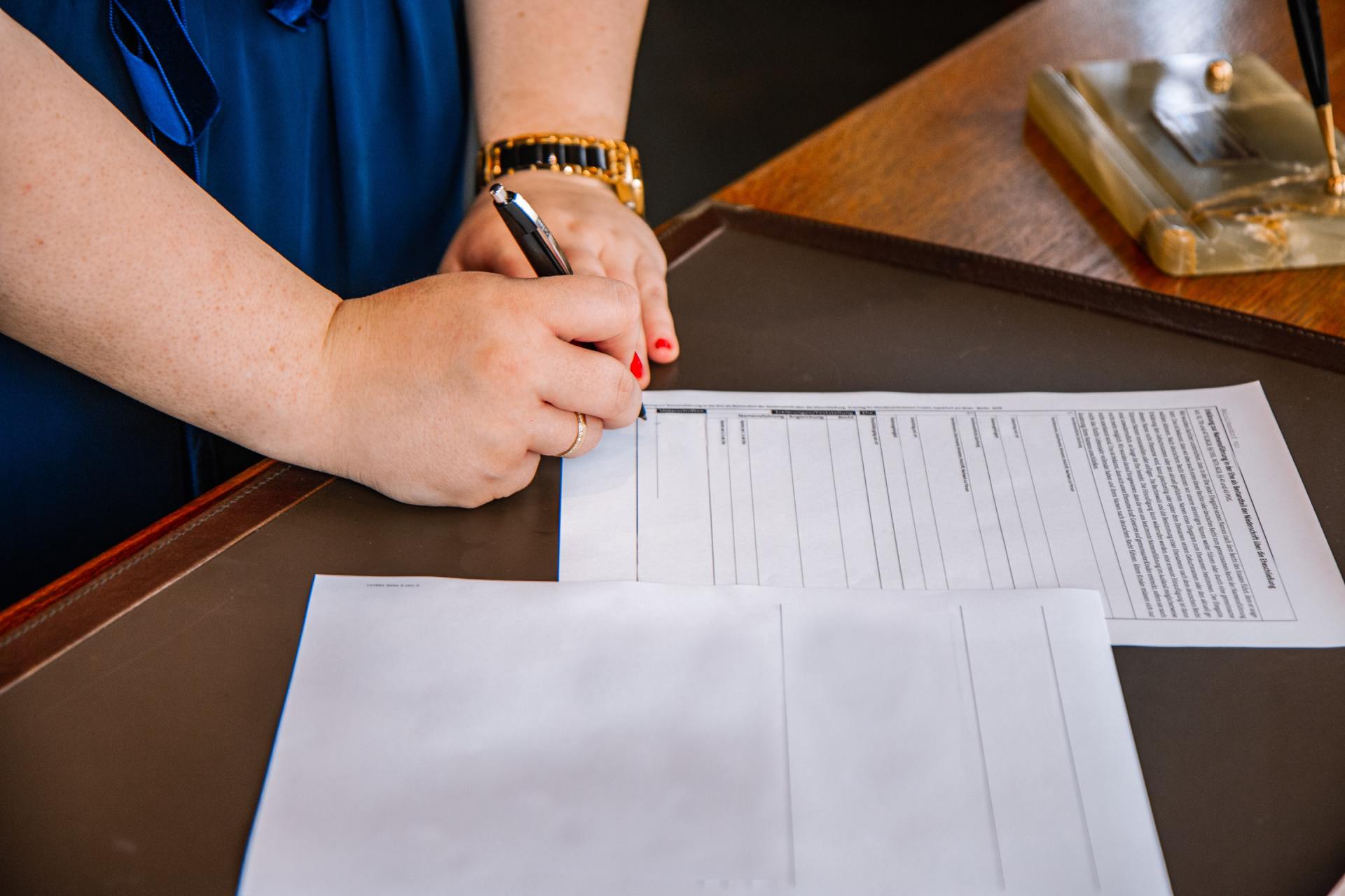 A person signing a document with a pen, with gold watch and red nail polish visible.