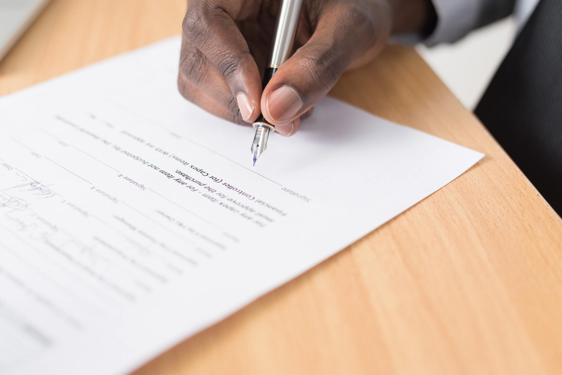 A person's hand signing a document with a silver pen on a wooden table.